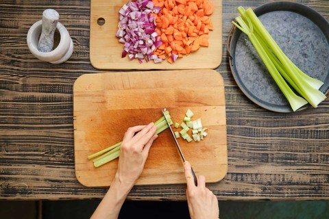 A person is chopping celery on a wooden cutting board, surrounded by diced onions and carrots. The