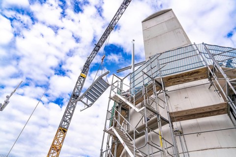 Construction crane lifts staircase structure next to tower and scaffolding in front of a cloudy
