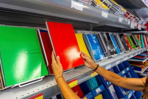 A person is holding a green spiral notebook in a store. There are many notebooks on the shelves,