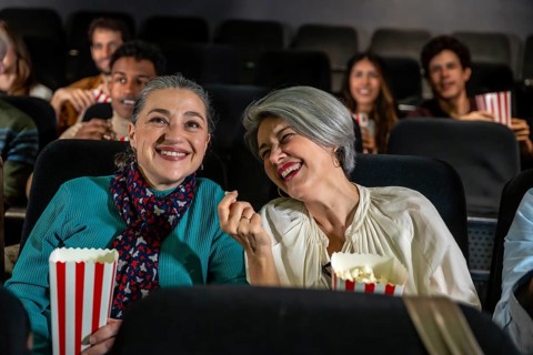 Two cheerful senior women laughing and eating popcorn while watching a movie at the cinema, sharing