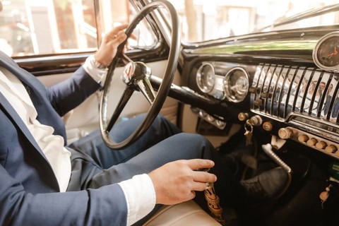 A man in a suit is driving a car with a black steering wheel. The car is a vintage model and the