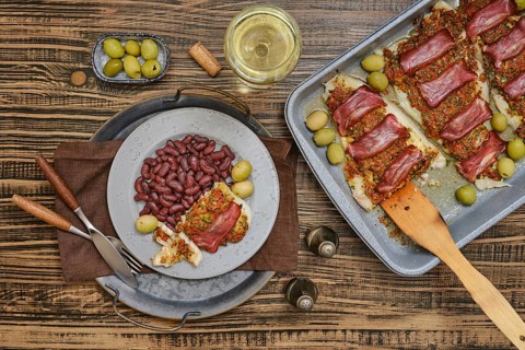 Baked fish topped with a crusty bacon, surrounded by green olives and red beans. A glass of white