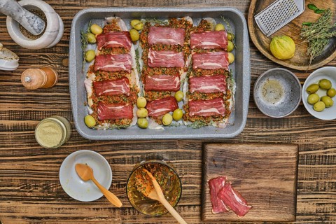 Fresh fish fillets layered with bacon and seasoning, herbs, and olives sits on a baking tray.