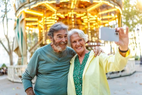 Cute smiling caucasian senior couple taking selfie in the city next to carousel