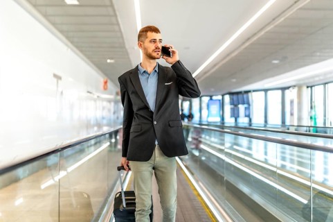 Businessman pulling suitcase and talking on smartphone while walking on moving walkway in airport