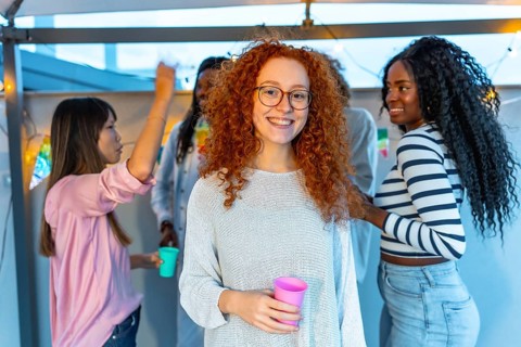 Young woman with redhead curly hair and eyeglasses smiling standing with beer glass in a roof party