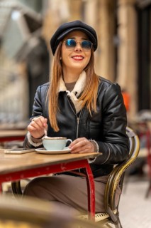 Young woman wearing sunglasses and baker boy hat stirring cappuccino at outdoor cafe table with