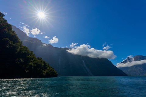 Sunbeams shine over majestic mountains and a calm lake under a clear sky, summer, Milford Sound, Te