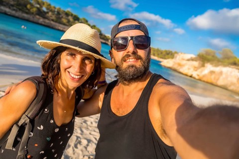 A man and woman are posing for a picture on a beach. The man is wearing a black tank top and