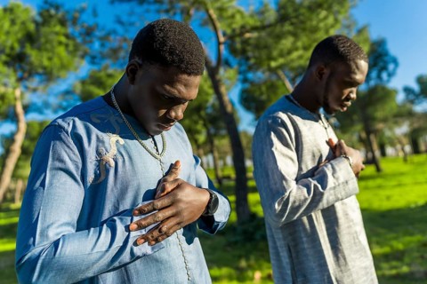 Two young senegalese men wearing traditional dashiki clothing are praying outdoors in a park,