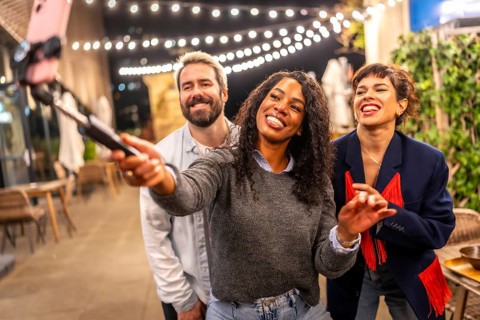 Latin woman streaming online during night party with multi-ethnic friends in the roof terrace of a