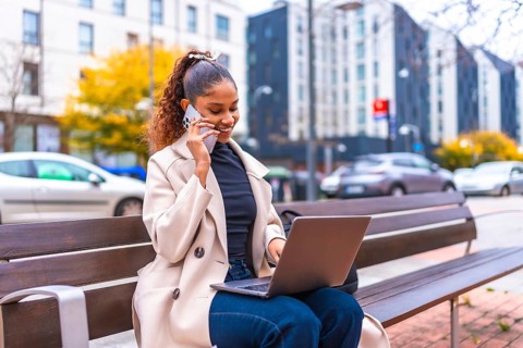 African businesswoman talking to the phone and using laptop sitting on a city bench