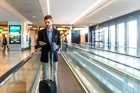 Businessman navigating airport terminal, using smartphone and enjoying coffee while walking on