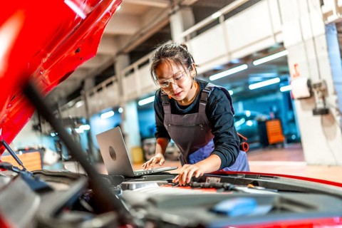 Young woman working as a mechanic, using a laptop while inspecting the engine of a red car in a