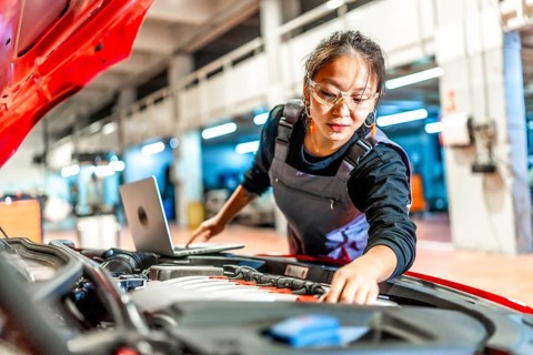 Young woman wearing mechanic overalls inspecting car engine using a laptop in a modern workshop