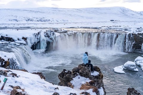 Woman hiker sitting looking at frozen and snowy Godafoss waterfall in winter in Iceland