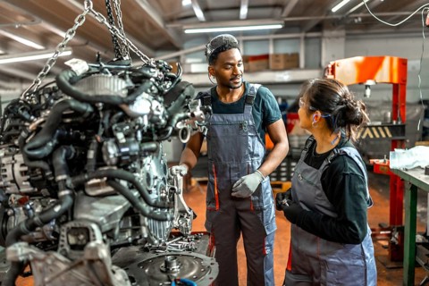Two auto mechanics, a man and a woman, examining a car engine suspended by chains in a repair shop,