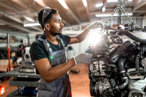 Young mechanic wearing protective gear working on a car engine suspended by a chain hoist in a