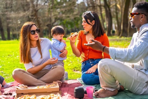 Family and friends enjoying pizza while picnicking in a vibrant green park, laughing and sharing