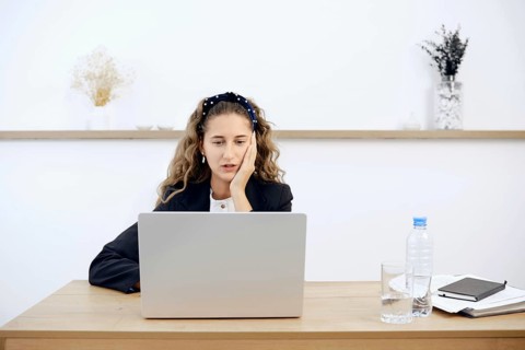 A young woman sits at a wooden desk, focused on her laptop while resting her chin on her hand. A
