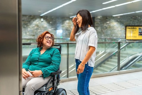 Smiling caucasian adult disabled woman and asian female friend talking waiting for subway elevator