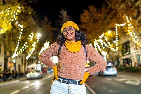 Young black woman tourist wearing yellow winter clothes and backpack, smiling with hands on her