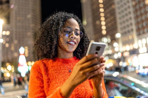 Young woman wearing an orange sweater, smiling while using a smartphone on a bustling city street,
