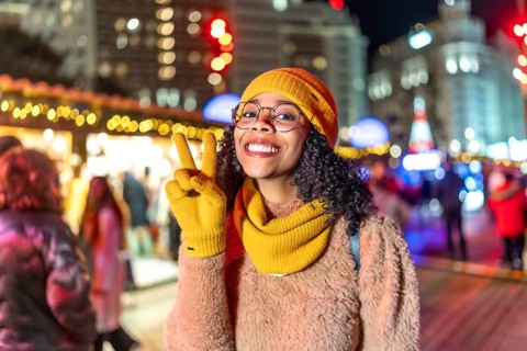 Smiling young woman wearing yellow gloves, hat and scarf showing peace sign while enjoying