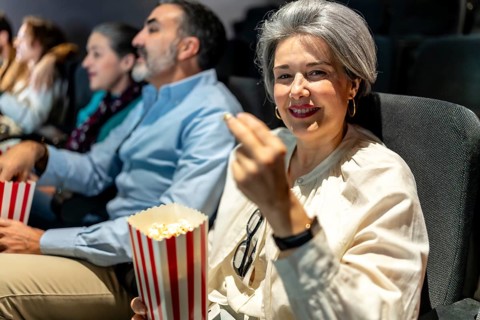 Mature woman smiling and eating popcorn while watching a movie at the cinema with friends, enjoying