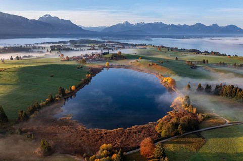 Aerial view of a small lake and autumn-coloured trees in front of mountains in the morning light,