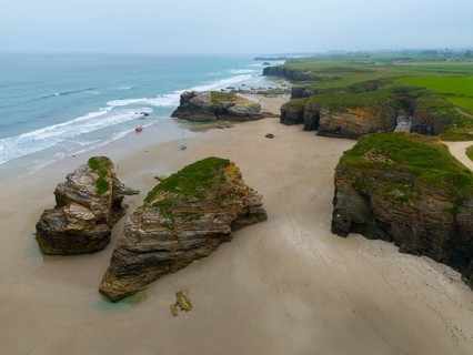 Wide beach with scattered large rock formations and calm water, bordered by steep coastal cliffs,