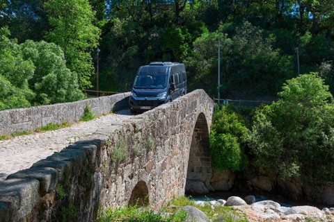 A van drives over a historic stone bridge, surrounded by lush vegetation and sunny weather,