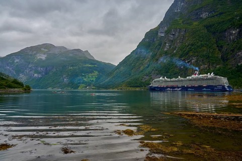 A large cruise ship, Mein Schiff 6, lies in the fjord, surrounded by green mountains under a cloudy