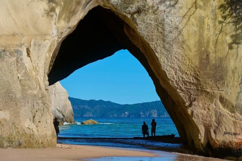 Silhouettes of people under a large rock arch on the beach overlooking the sea, Cathedral Cove,