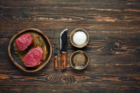 Two raw steaks on a wooden plate alongside bowls of salt and pepper, with old forged knife and fork