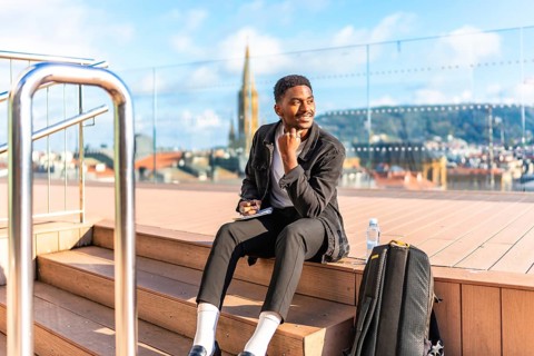Happy african man sketching by hand in a notebook sitting on a rooftop with city views
