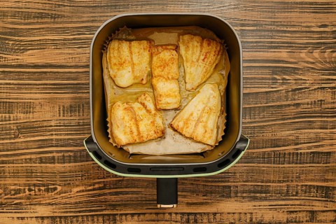 Five golden brown grilled fish fillets are neatly placed inside an air fryer basket. The background