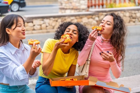 Hungry female multiracial friends eating take away pizza sitting outdoors in the city