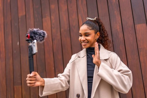 Young african american woman influencer waving at camera while recording vlog outdoors next to a