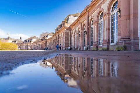 Large building with elaborate architecture reflected in a puddle under a blue sky, Schwetzingen