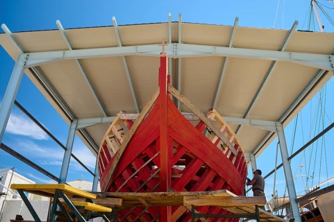 Front view of a partially built red wooden ship in a covered shipyard under a clear sky, Patmos