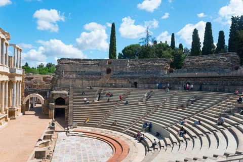 An ancient amphitheatre with stone seats under a blue sky with clouds and trees in the background,