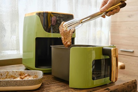 A hand holds tongs putting a piece of marinated meat to an air fryer. The process of preparing