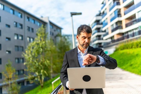 Mature adult caucasian businessman checking time to smartwatch while standing using laptop at city