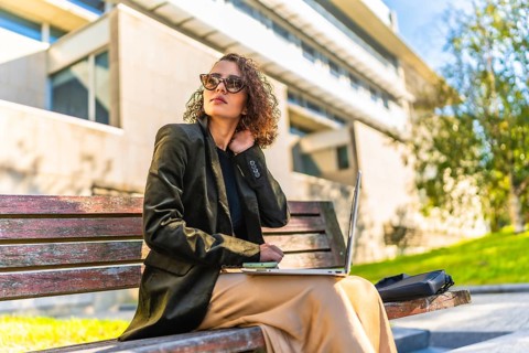 Young arab female freelancer working with laptop sitting on park bench under the sun