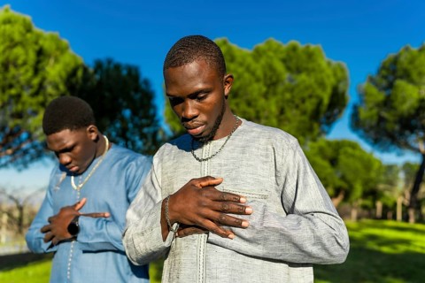 Two young senegalese men wearing traditional clothing are praying outdoors in a park, expressing
