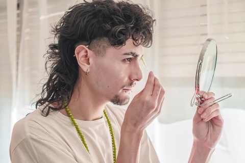 Concentrated young man with curly hair plucking his eyebrows with tweezers while looking in a small