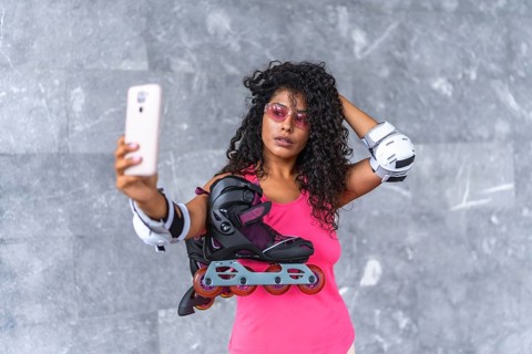 Latin woman in sportswear and protective gear taking selfie holding inline skates outdoors