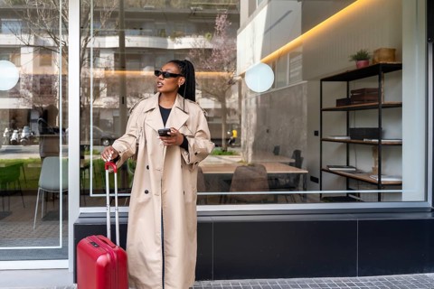 Stylish businesswoman holding smartphone and pulling rolling luggage while arriving at a hotel on a