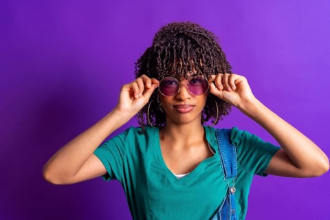 Stylish young woman lowering her purple sunglasses while posing in a vibrant purple studio,